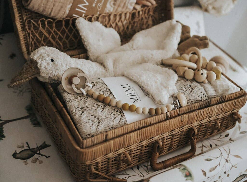 Wicker basket with baby items including a plush toy, blanket, and wooden beads on a floral-patterned surface.