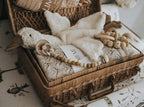 Wicker basket with baby items including a plush toy, blanket, and wooden beads on a floral-patterned surface.